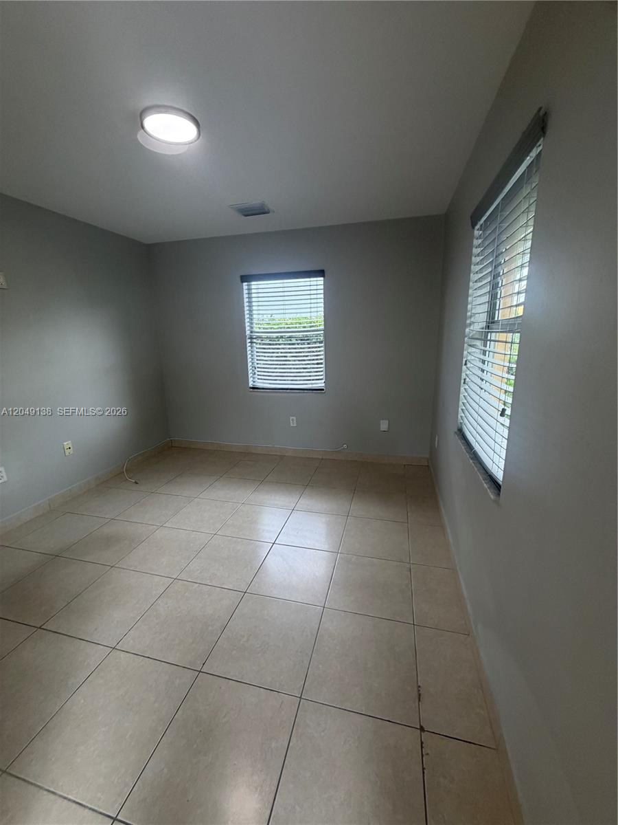 An empty room features light-colored tile flooring, gray walls, and two windows with blinds — PALMETTO BAY, FL