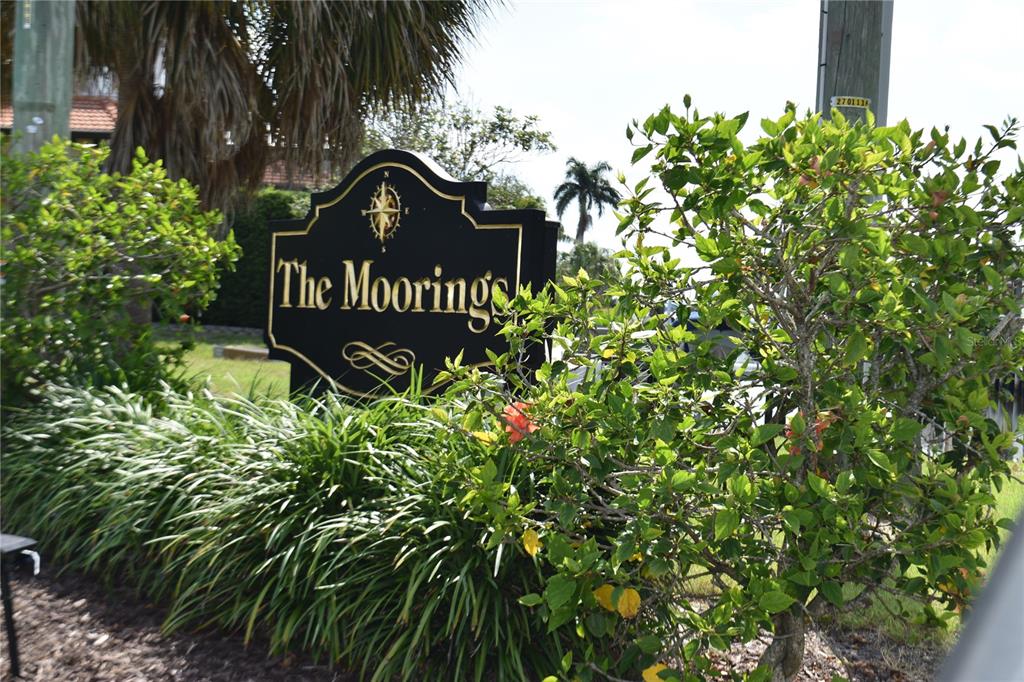 A black sign with gold lettering reading The Moorings stands among green bushes and trees — 420 MOORINGS COVE DRIVE UNIT 420, TARPON SPRINGS, FL