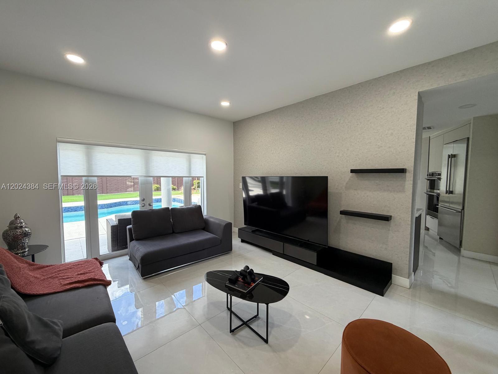 A living room features a large television, dark furniture, and a view of a pool — 10462 NW 130th St # 1, Hialeah Gardens FL 33018, HIALEAH GARDENS, FL