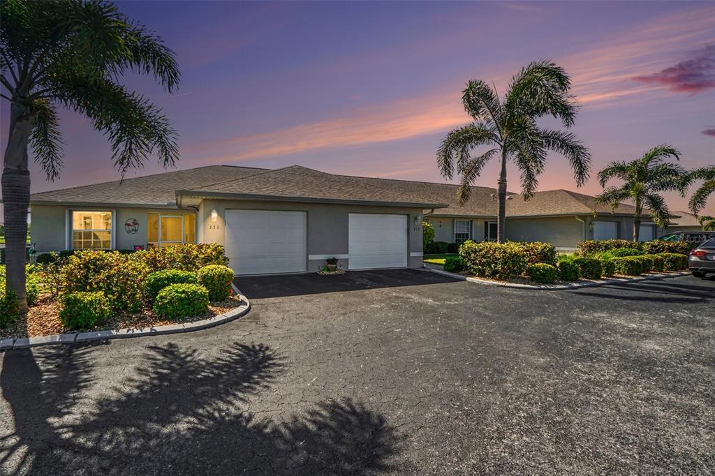 A single-story duplex with two garage doors, palm trees, and landscaping under a sunset sky — 26333 NADIR ROAD UNIT 111, PUNTA GORDA, FL
