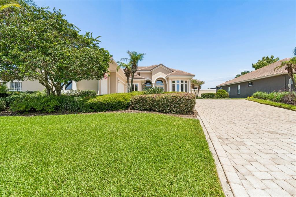 A beige house with a tiled roof and a paved driveway sits behind a lawn — 12749 GRAND TRAVERSE DRIVE, DADE CITY, FL