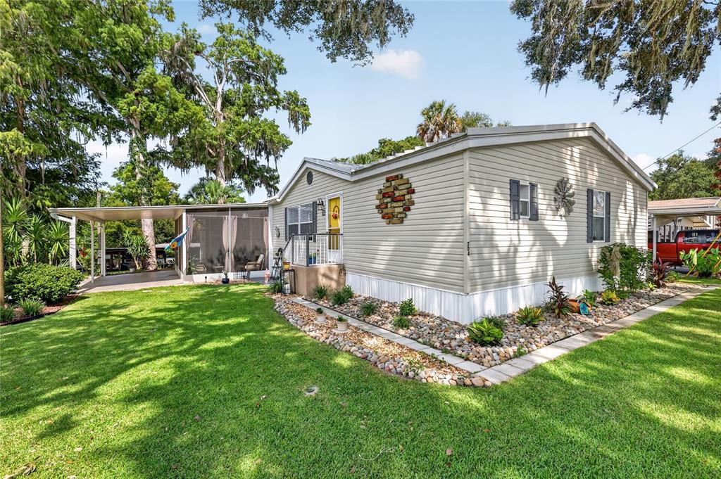 A light-colored mobile home with a screened-in porch, rock landscaping, and a large grassy yard — 704 KIMBERLY AVENUE, LEESBURG, FL