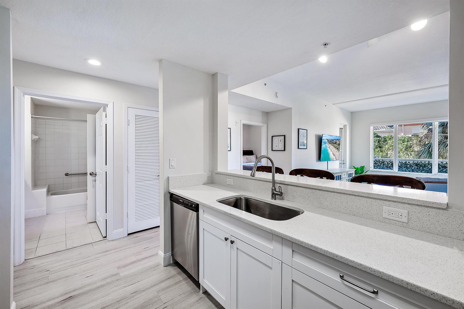 A kitchen with white cabinets, quartz countertops, and a stainless steel sink overlooks a living area — 1203 Town Center Drive # 201, Jupiter FL 33458, JUPITER, FL
