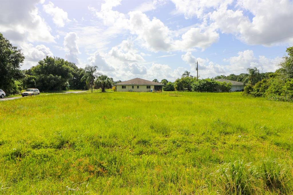 A large, grassy vacant lot sits in front of a single-story house under a cloudy sky — 12000 TAMIAMI LOTS 15 & 16 TRAIL S, NORTH PORT, FL