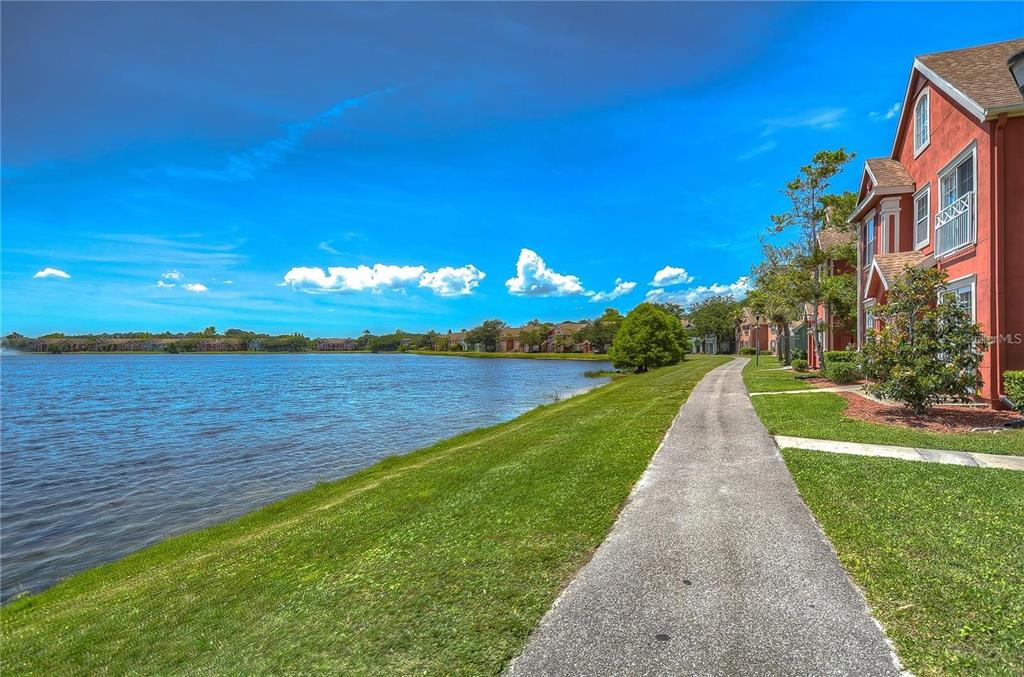 A paved walkway runs along a grassy bank beside a lake next to red buildings — 9034 LAKE CHASE ISLAND WAY UNIT 9034, TAMPA, FL