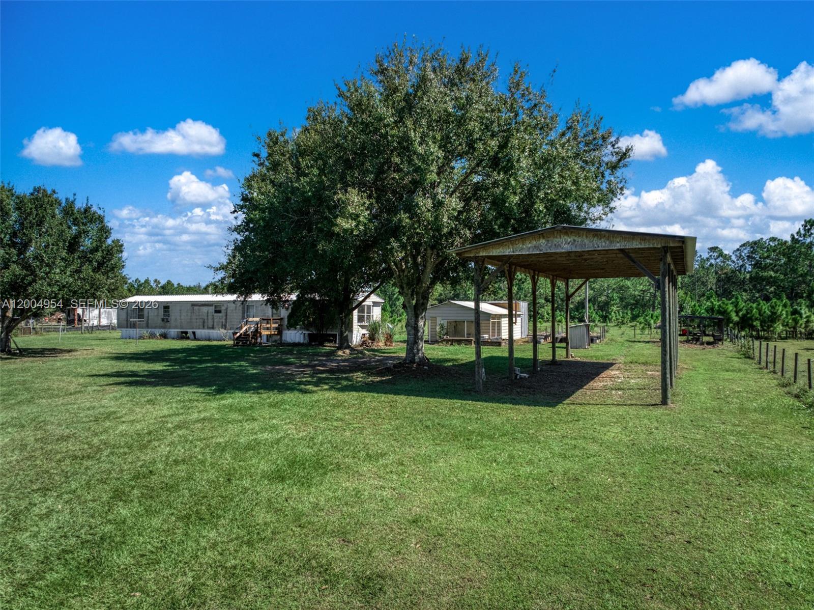 A grassy yard features a mobile home, a large tree, and a wooden carport structure — 2316 Gem Stone, Sebring FL 33857, SEBRING, FL