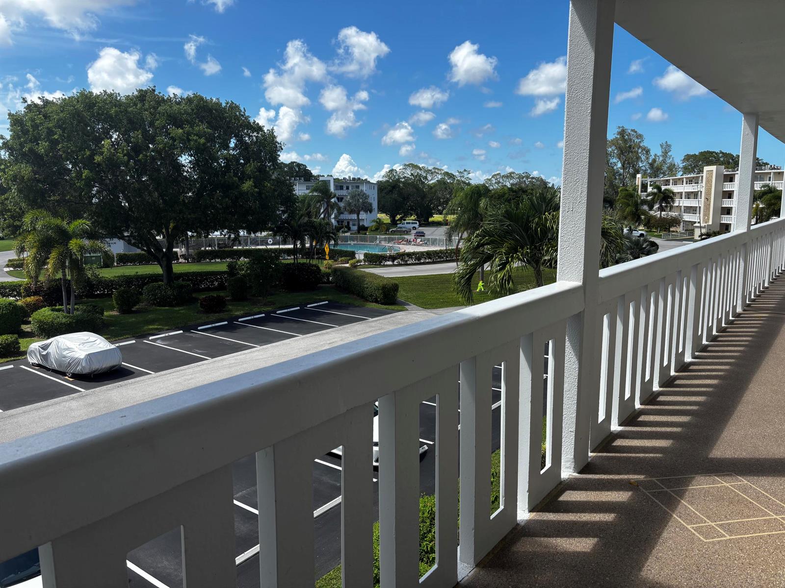 A view from a white balcony overlooking a parking lot, palm trees, and a pool — 309 Grantham A, Deerfield Beach FL 33442, DEERFIELD BEACH, FL