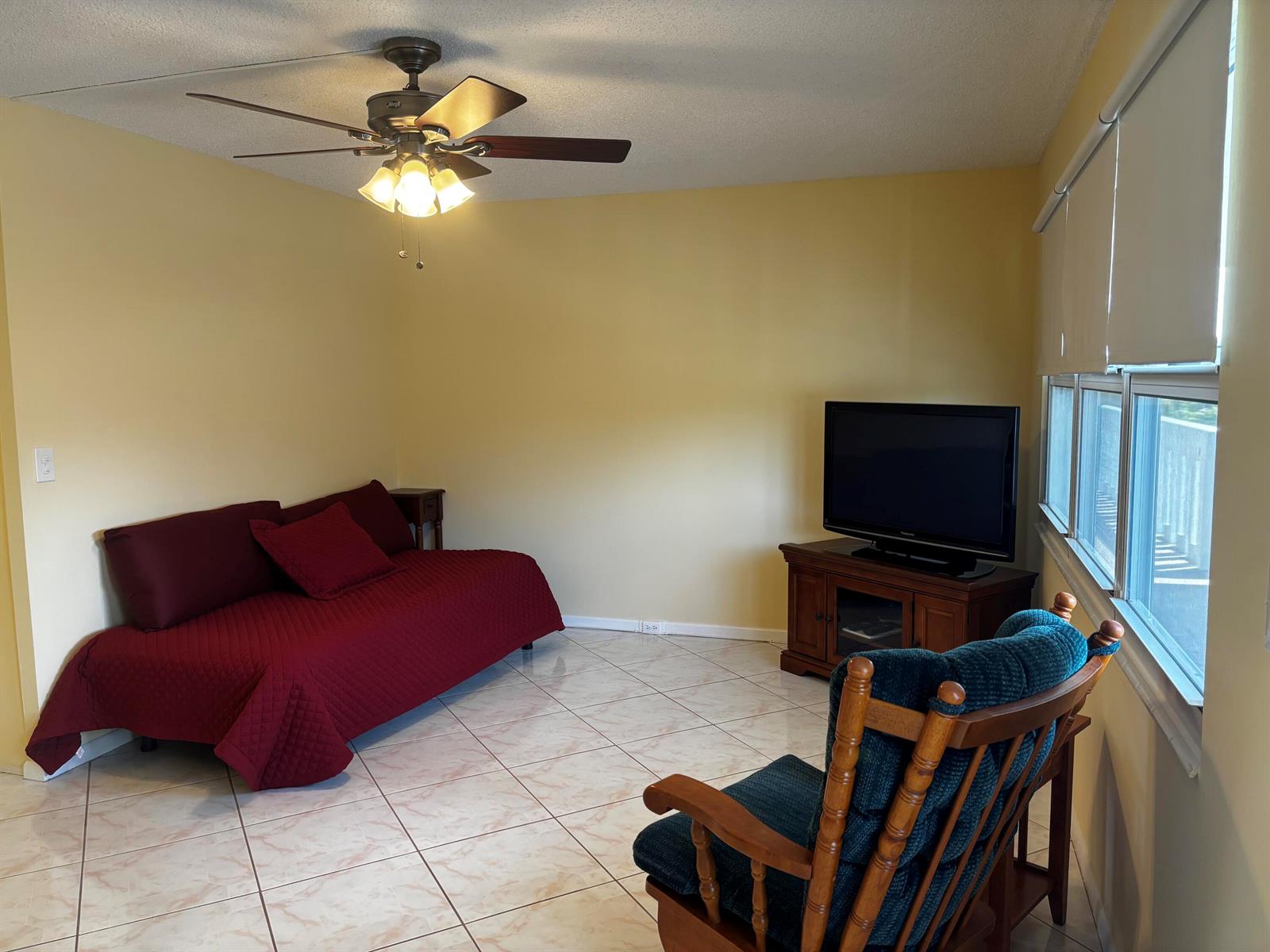 A living room with yellow walls, tile flooring, a daybed, a television, and a rocking chair — 309 Grantham A, Deerfield Beach FL 33442, DEERFIELD BEACH, FL