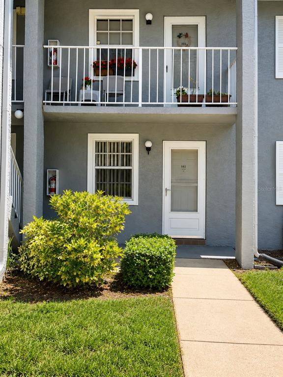 A two-story apartment building exterior features a ground-level entrance, a balcony, and landscaping — 1420 OAK HILL DRIVE UNIT 102, DUNEDIN, FL