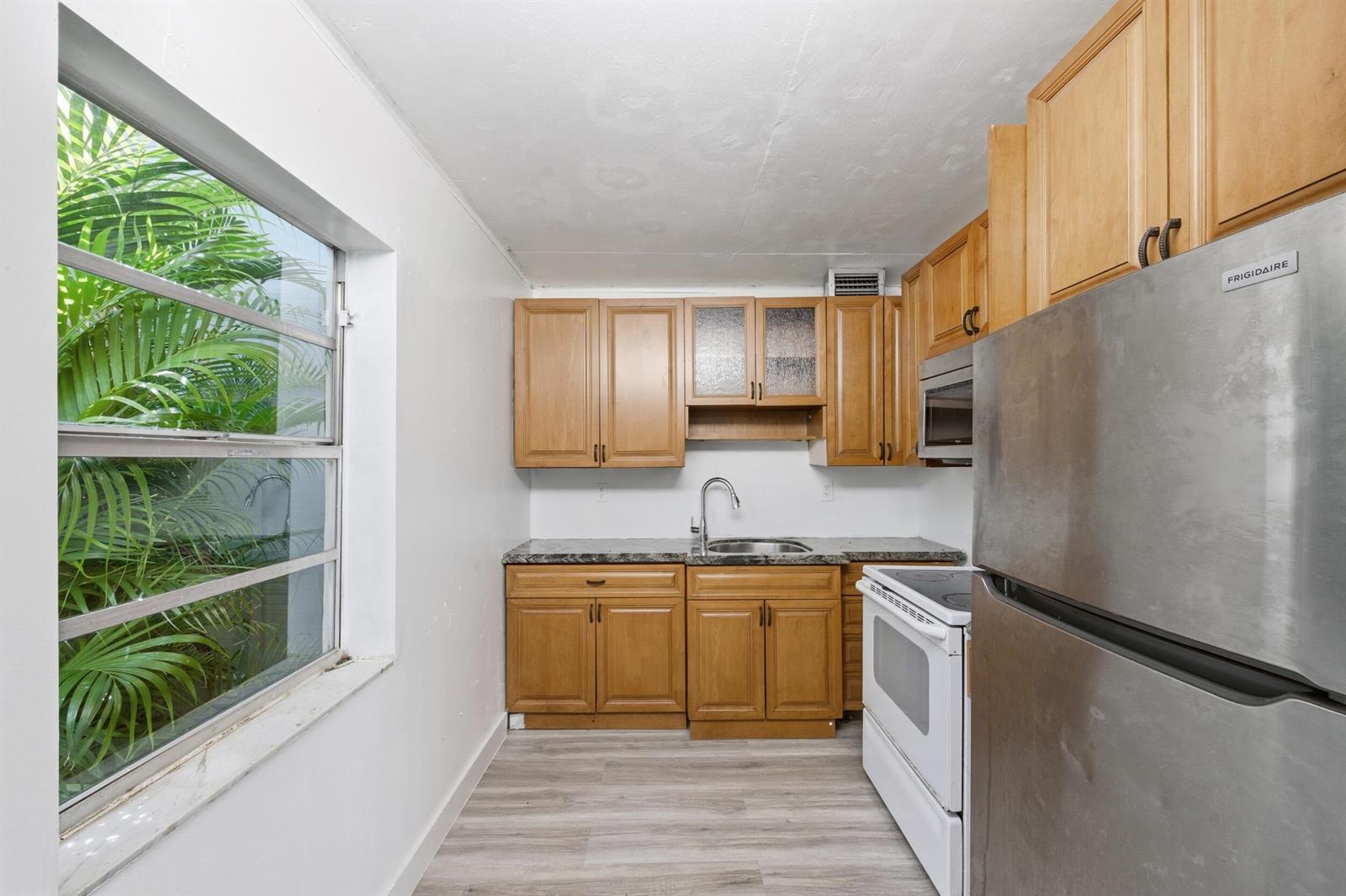 A small kitchen features wood cabinets, stainless steel appliances, and a window overlooking palm trees — 452 Normandy # J, Delray Beach FL 33484, DELRAY BEACH, FL