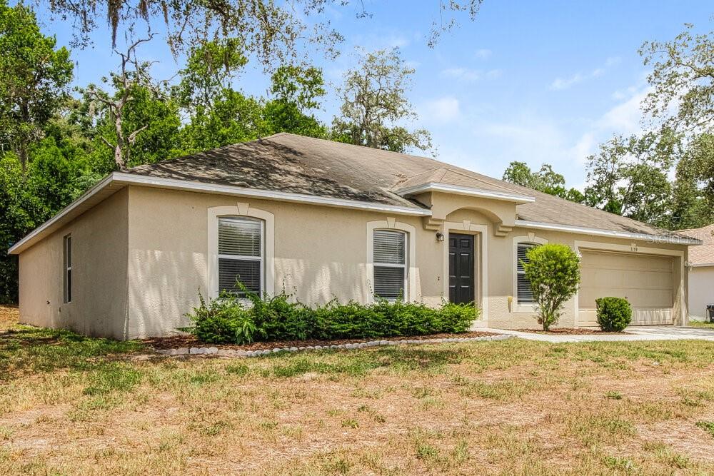A single-story tan stucco house with a dark front door and a two-car garage — 1159 NEWCOMB AVENUE, SPRING HILL, FL
