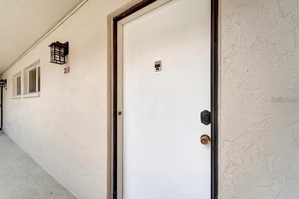A white exterior door with a peephole and two locks is labeled with the number 211 — 2763 WOODGATE LANE UNIT 211, SARASOTA, FL