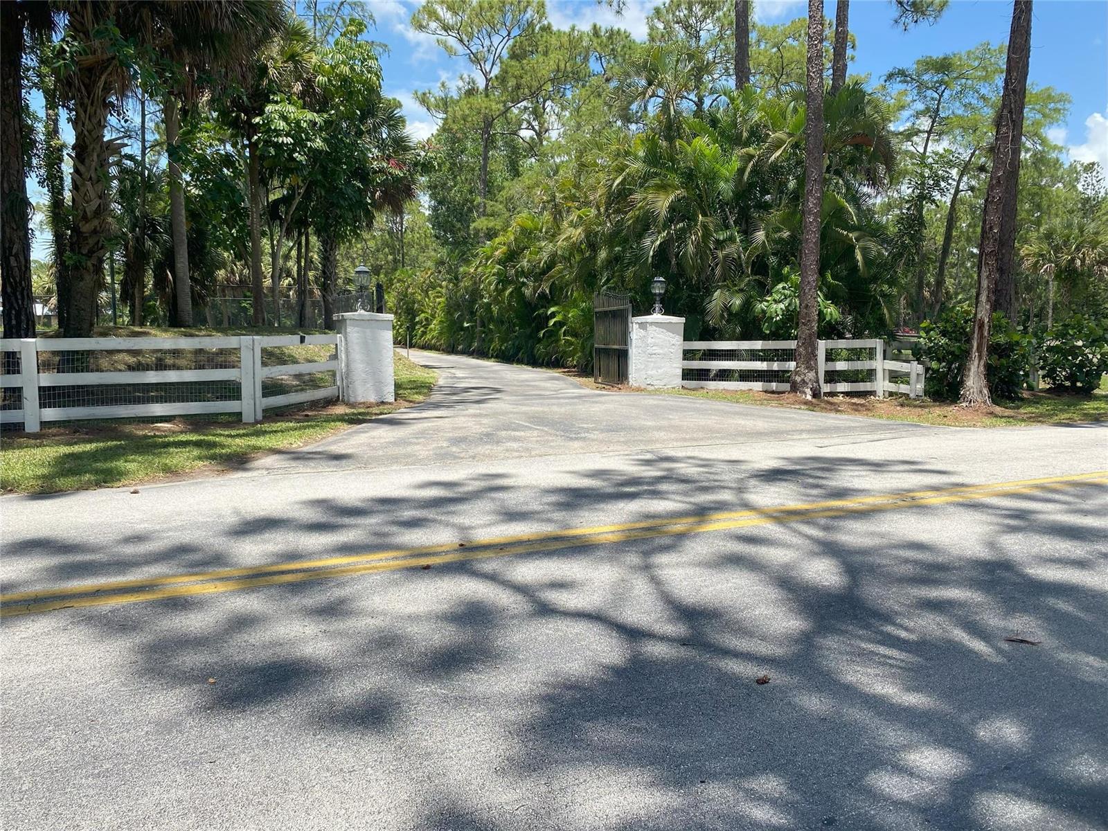 A paved driveway leads through a gated entrance with white fencing and lush landscaping — 2315 Deer Run Boulevard, Loxahatchee FL 33470, LOXAHATCHEE, FL