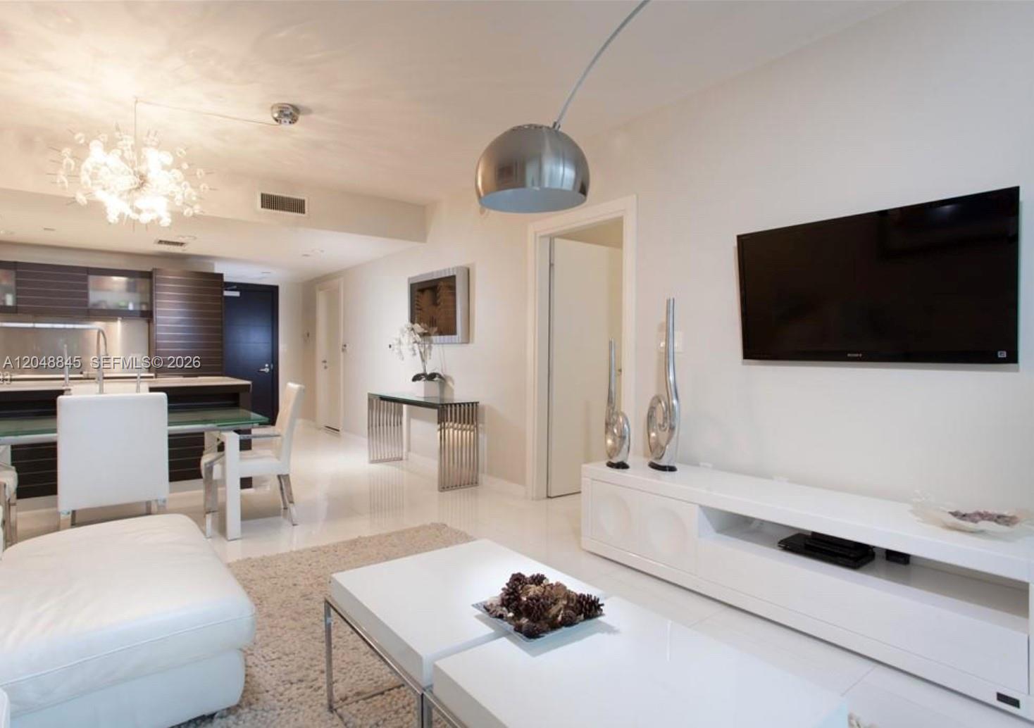 A modern living room features white furniture, a glass coffee table, and a large chandelier — 200 Biscayne Boulevard Way # 3209, Miami FL 33131, MIAMI, FL