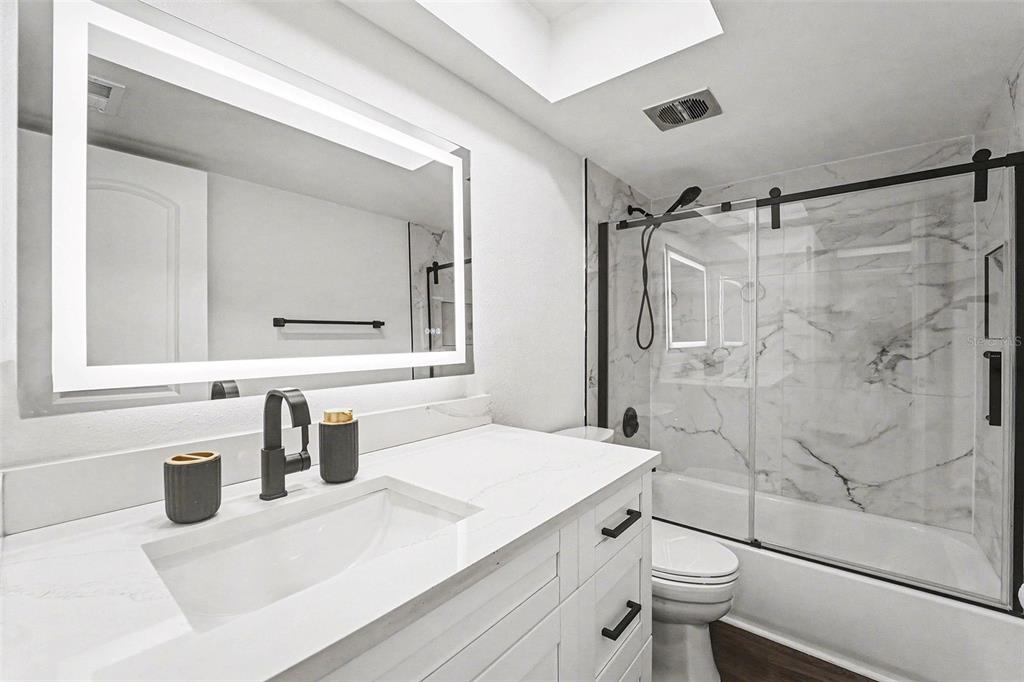 A modern bathroom features a white vanity, a backlit mirror, and a marble-tiled shower-tub combo — 5197 SILENT LOOP UNIT 121, NEW PORT RICHEY, FL