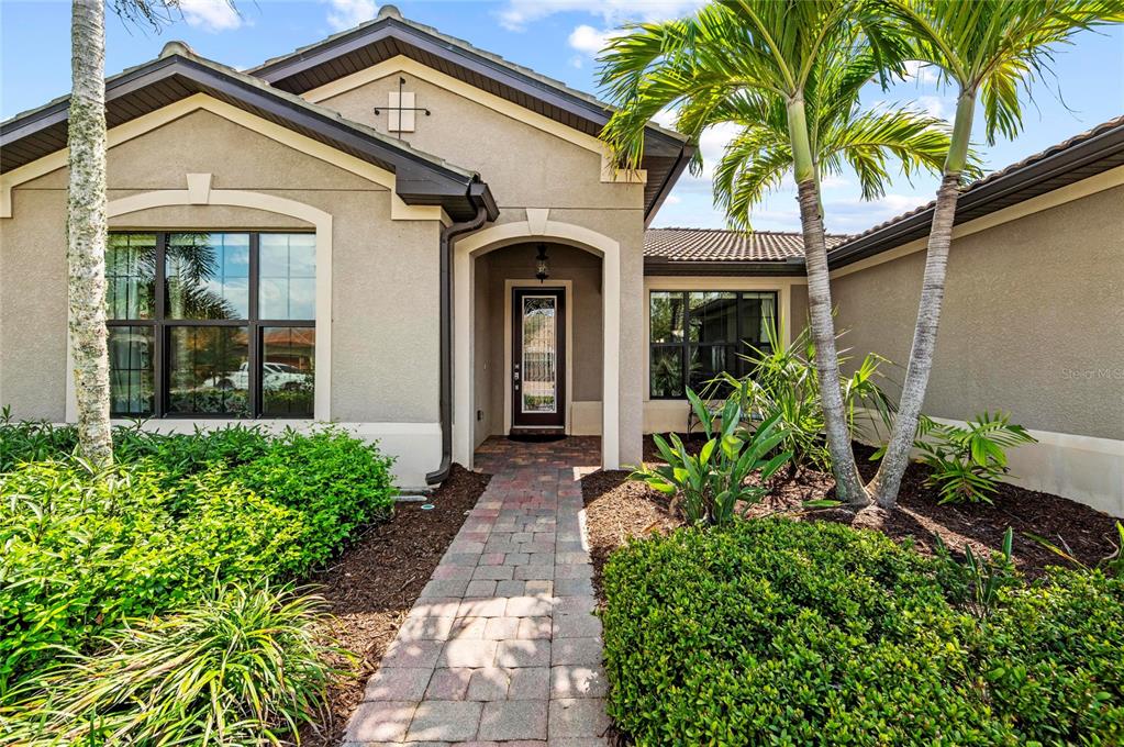 A paved walkway leads to the front entrance of a tan stucco house with palm trees — 13785 TOMARO STREET, VENICE, FL
