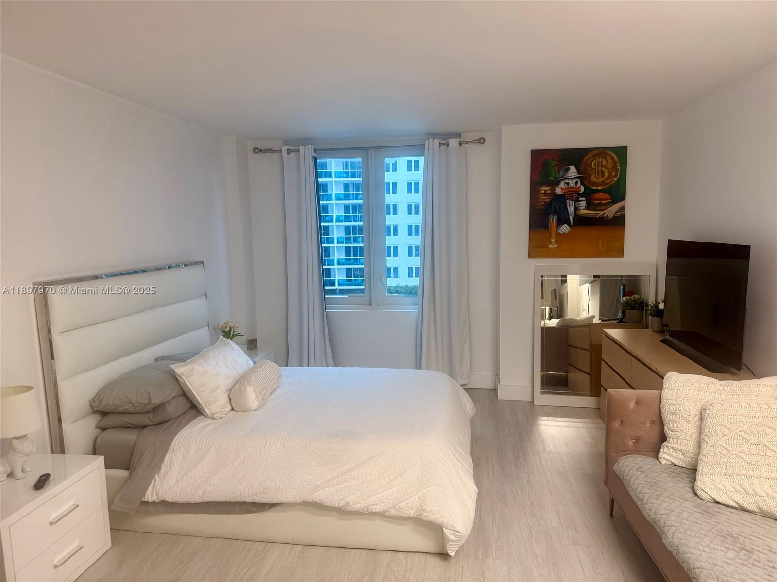 A bedroom features a white upholstered bed, light wood flooring, and a wall-mounted television — 2301 Collins Ave # 619, Miami Beach FL 33139, MIAMI BEACH, FL