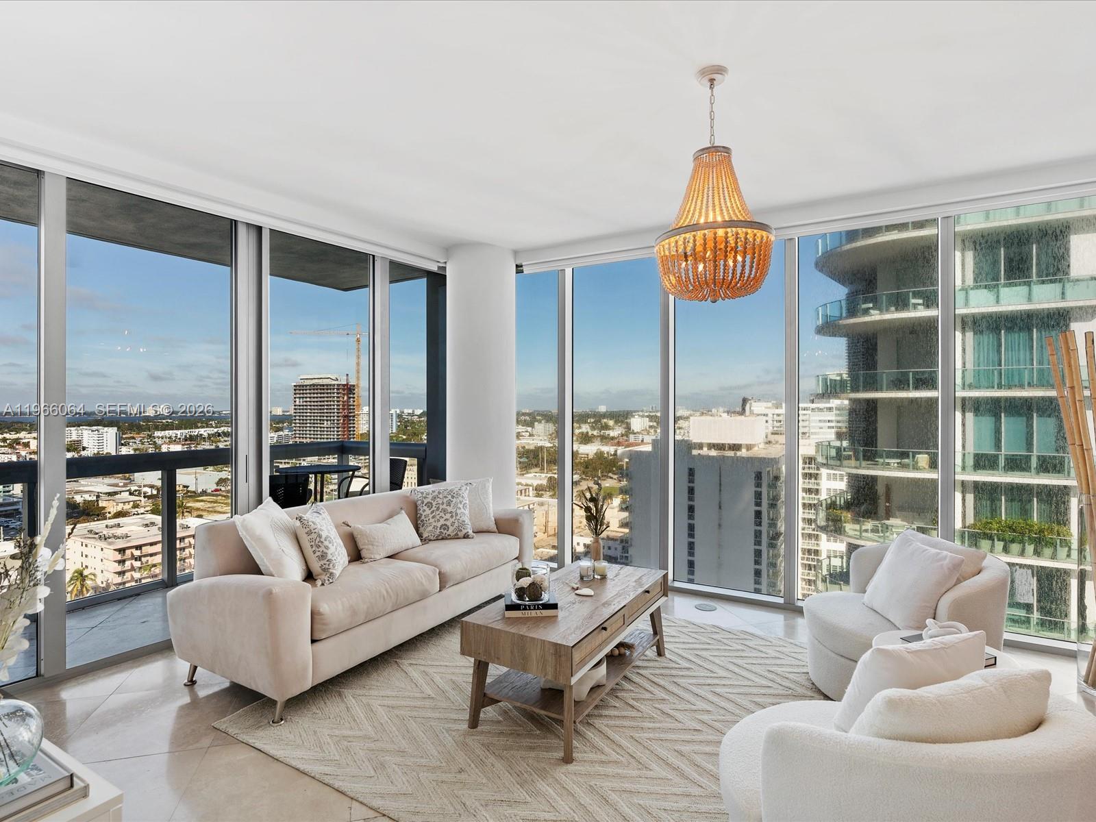 A modern living room features floor-to-ceiling windows, a beige sofa, and a wooden chandelier — 6899 Collins Ave # 1704, Miami Beach FL 33141, MIAMI BEACH, FL