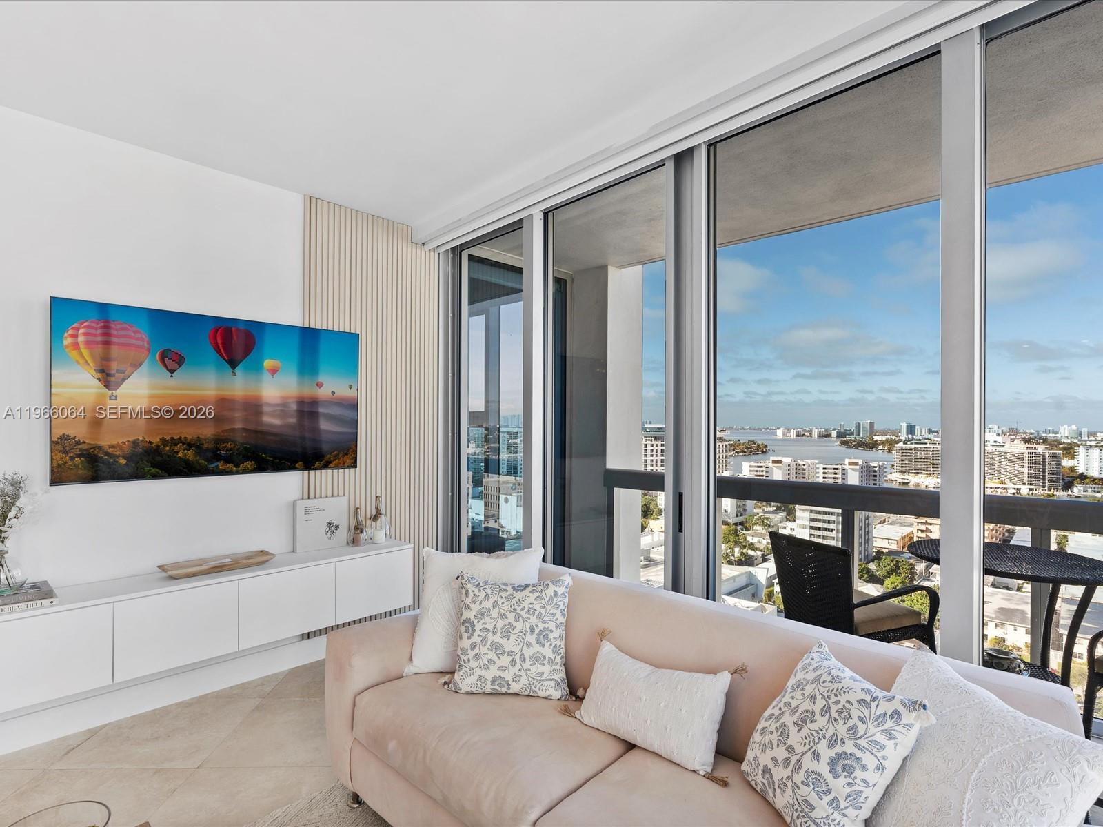 A living room features a wall-mounted television, a light-colored sofa, and floor-to-ceiling windows — 6899 Collins Ave # 1704, Miami Beach FL 33141, MIAMI BEACH, FL