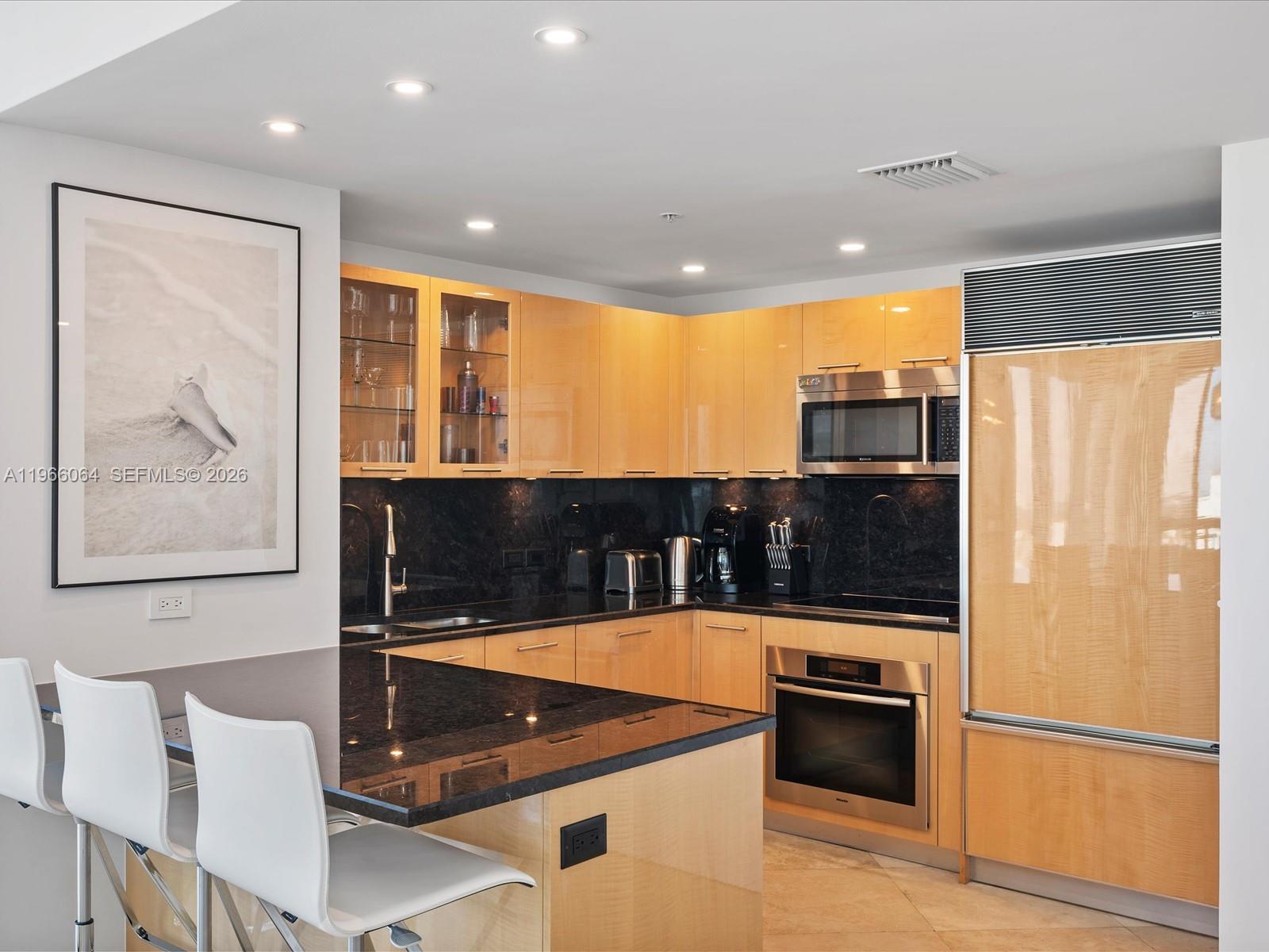 A modern kitchen features light wood cabinets, black granite countertops, and stainless steel appliances — 6899 Collins Ave # 1704, Miami Beach FL 33141, MIAMI BEACH, FL