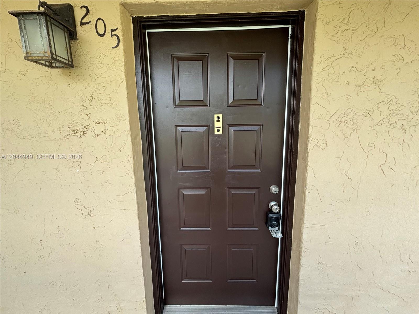 A brown six-panel door with a lockbox is set into a textured beige exterior wall — 21909 Lake Forest Cir # 205, Boca Raton FL 33433, BOCA RATON, FL