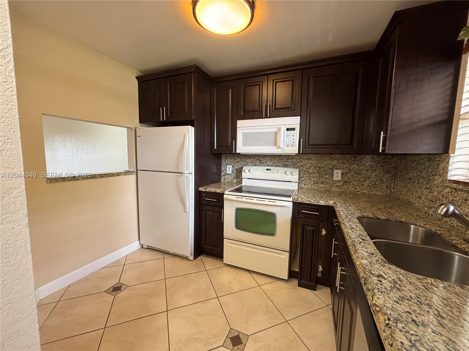 A kitchen features dark wood cabinets, granite countertops, white appliances, and beige tile flooring — 21909 Lake Forest Cir # 205, Boca Raton FL 33433, BOCA RATON, FL