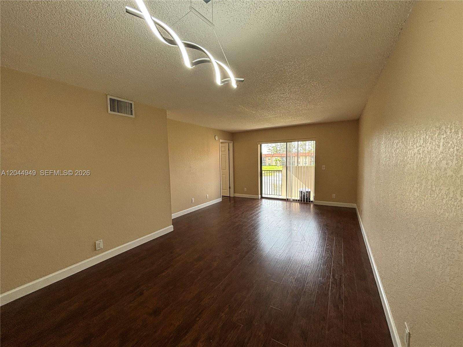 An empty living room features dark wood-look flooring, beige walls, and a modern light fixture — 21909 Lake Forest Cir # 205, Boca Raton FL 33433, BOCA RATON, FL