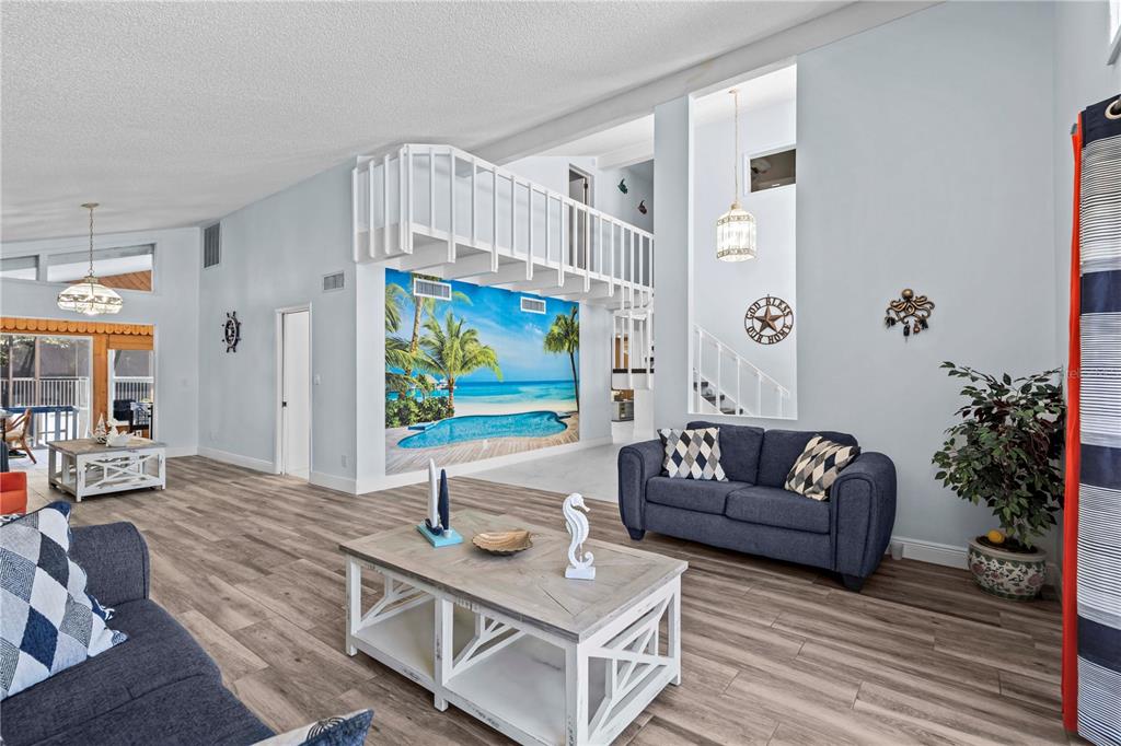 A living room features a large tropical wall mural, a lofted balcony, and wood-look flooring — 107 CAUSEWAY BOULEVARD, BELLEAIR BEACH, FL