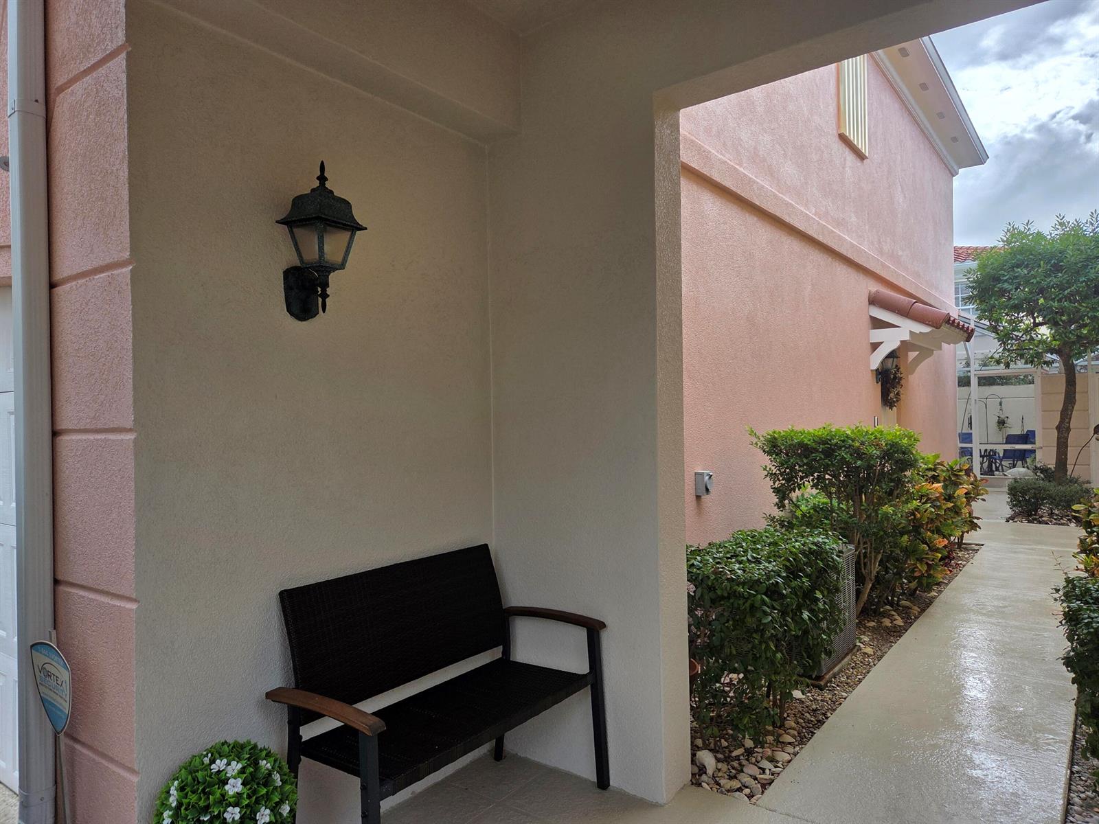 A covered entryway features a black bench, a wall-mounted light fixture, and exterior stucco walls — 58 Tall Oaks Circle, Tequesta FL 33469, TEQUESTA, FL