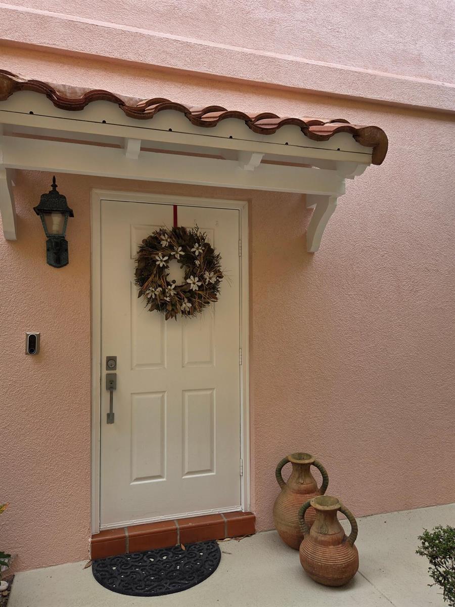 A white front door with a wreath sits under a tiled awning on a pink wall — 58 Tall Oaks Circle, Tequesta FL 33469, TEQUESTA, FL
