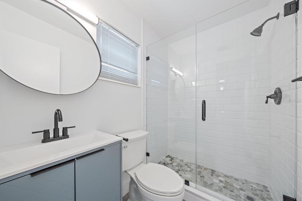 A bathroom features a blue vanity, a round mirror, a toilet, and a glass-enclosed shower — 3818 ROXANE BOULEVARD UNIT 157, SARASOTA, FL