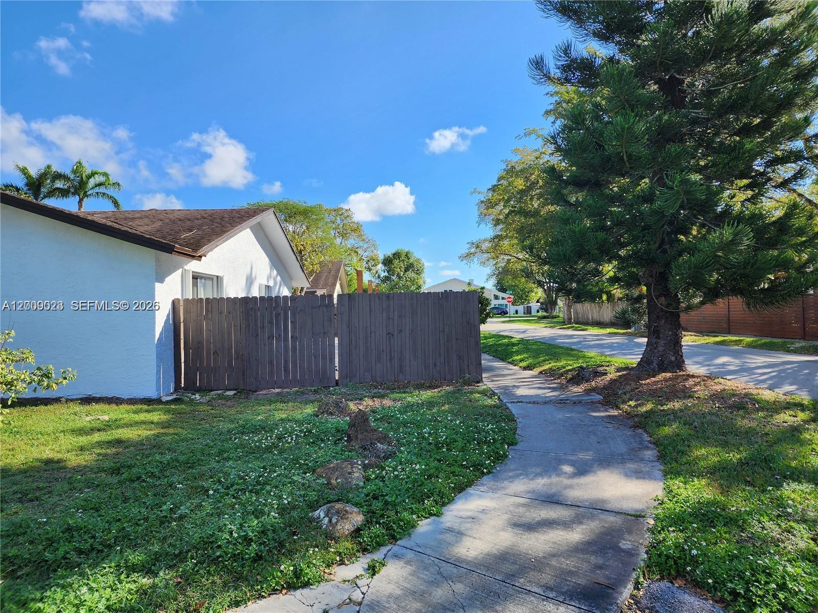 A white house with a brown roof sits behind a wooden fence near a sidewalk — 9825 SW 132nd Ct, Miami FL 33186, MIAMI, FL