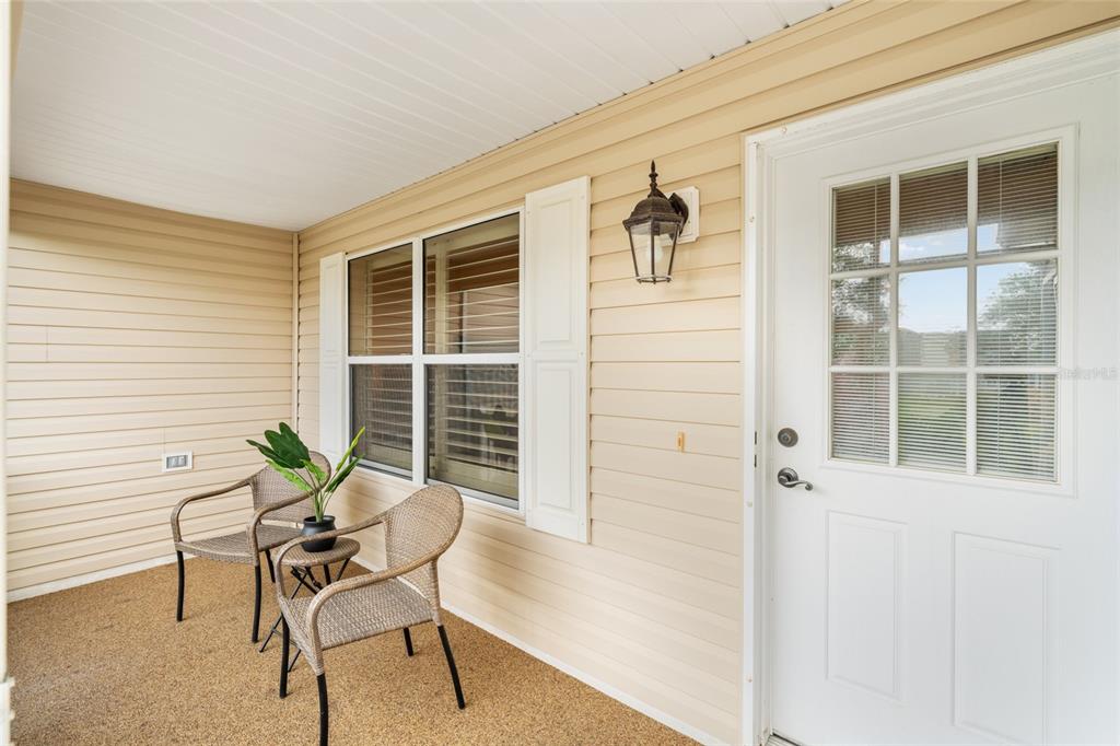 A covered porch features beige vinyl siding, two wicker chairs, and a white front door — 991 BARRYMOORE LOOP, THE VILLAGES, FL