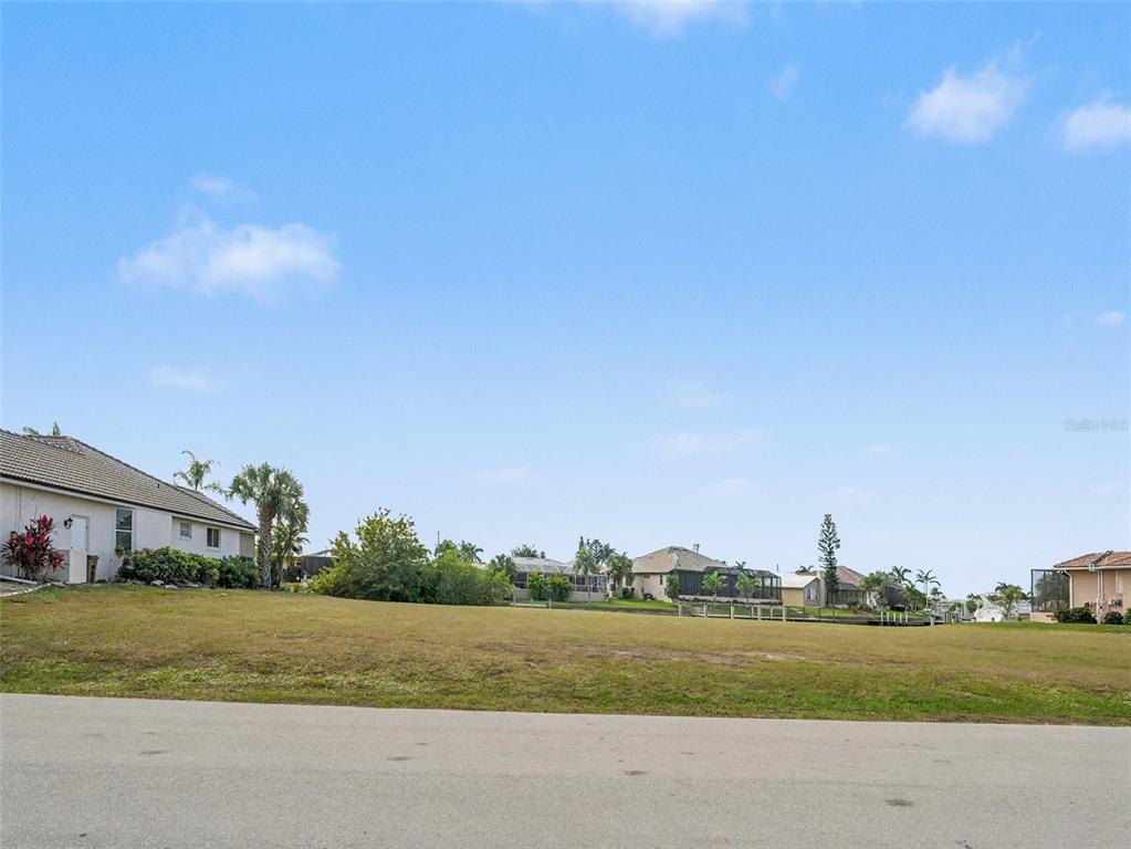 A grassy vacant lot sits along a paved road with neighboring houses in the background — 1247 PINE SISKIN DRIVE, PUNTA GORDA, FL