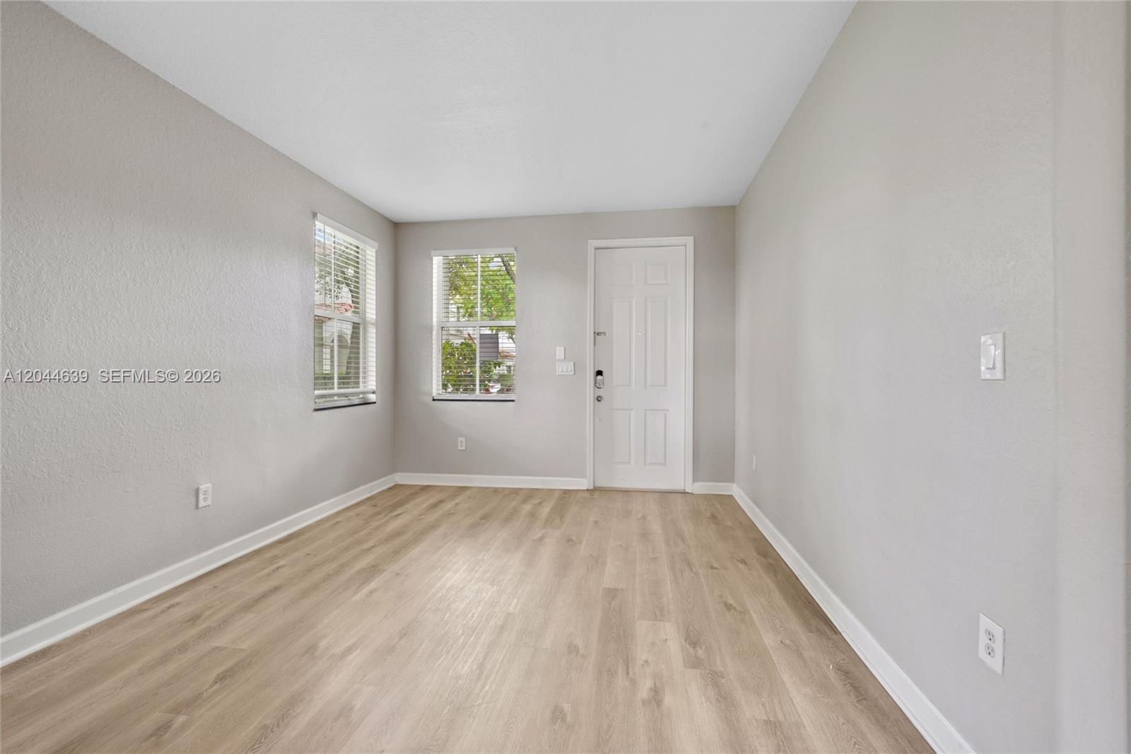 An empty room features light-colored wood-look flooring, gray walls, two windows, and a white door — 10709 NW 81st Ln, Doral FL 33178, DORAL, FL