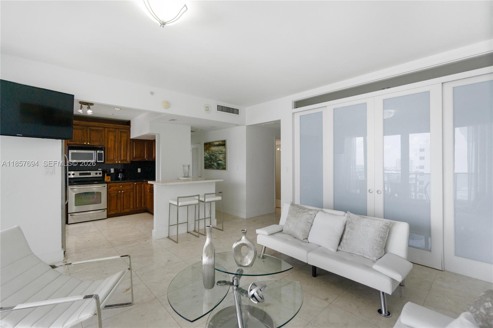 A living room with a white sofa, glass coffee table, and an adjacent kitchen area — 100 Lincoln Rd, Miami Beach FL 33139, MIAMI BEACH, FL