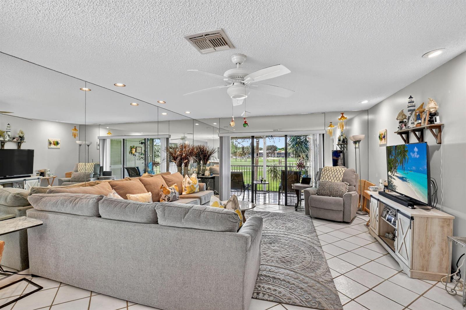 A living room features a large sectional sofa, mirrored wall, tile floors, and a balcony view — 7 Southport Lane # B, Boynton Beach FL 33436, BOYNTON BEACH, FL