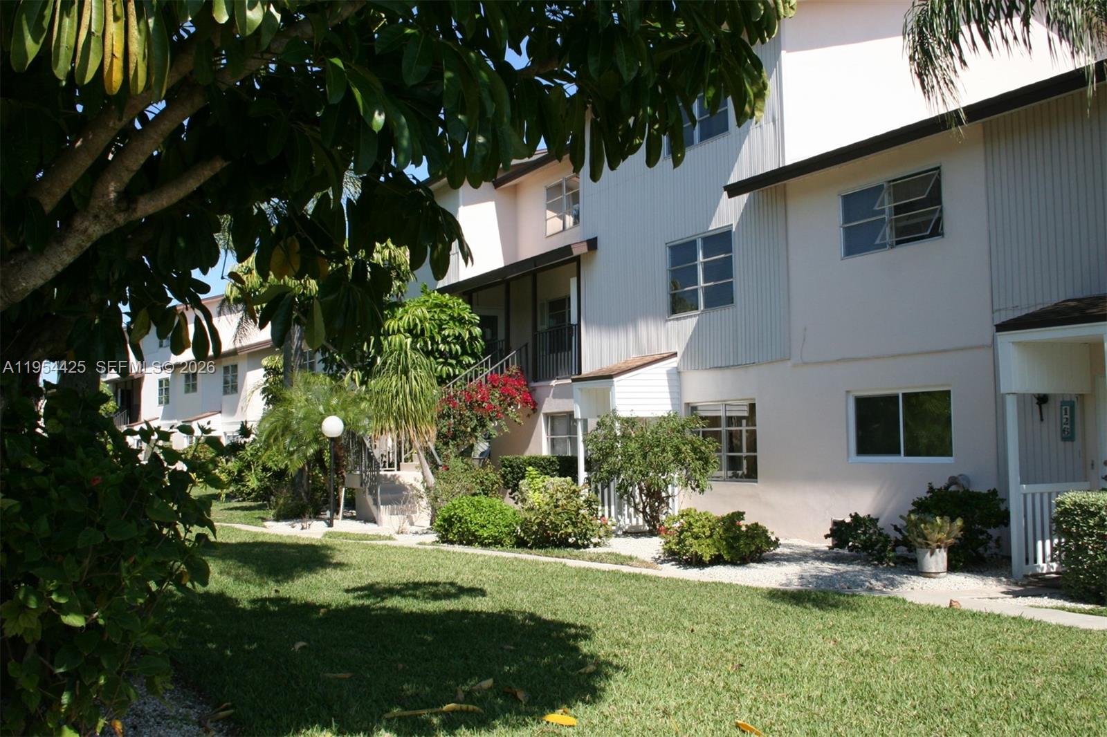 A two-story apartment building with light-colored siding, a grassy lawn, and lush landscaping — 180 E Cypress Way # E230, Naples FL 34110, NAPLES, FL