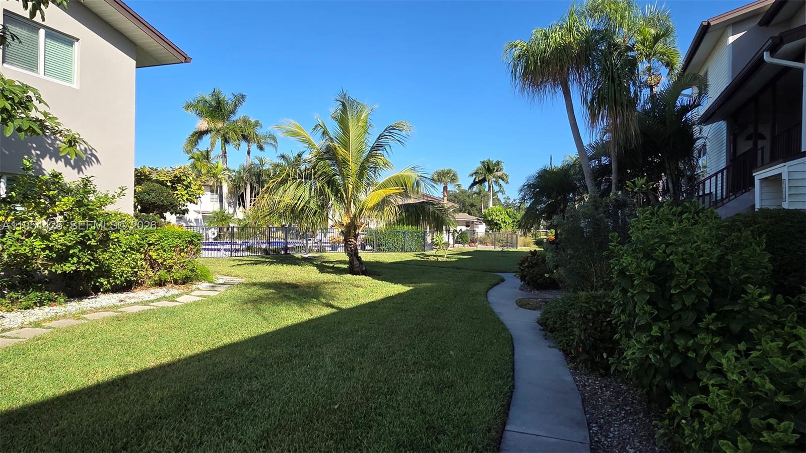 A grassy courtyard with a concrete walkway, palm trees, and a swimming pool in the background — 180 E Cypress Way # E230, Naples FL 34110, NAPLES, FL