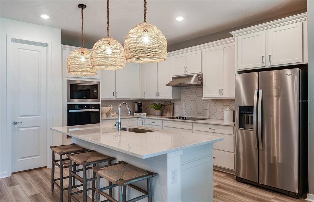 A modern kitchen features white cabinetry, a quartz-topped island with stools, and woven pendant lights — 37385 GUNNISON DRIVE, ZEPHYRHILLS, FL