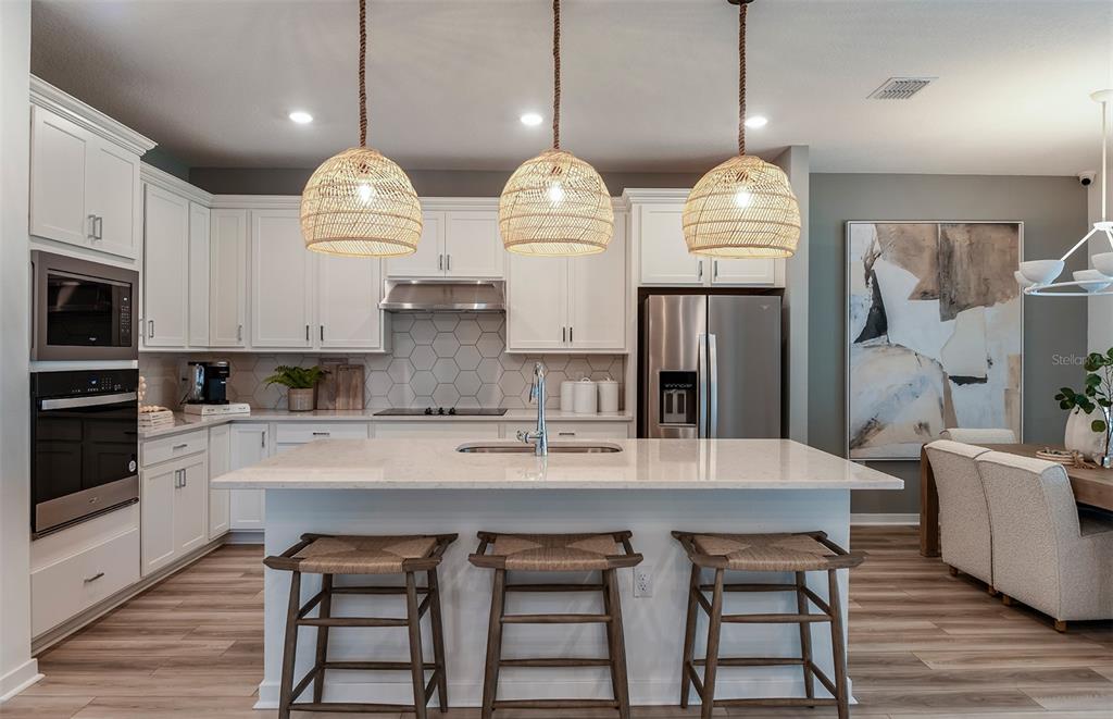 A modern kitchen features white cabinets, a quartz island with three stools, and woven pendant lights — 37385 GUNNISON DRIVE, ZEPHYRHILLS, FL