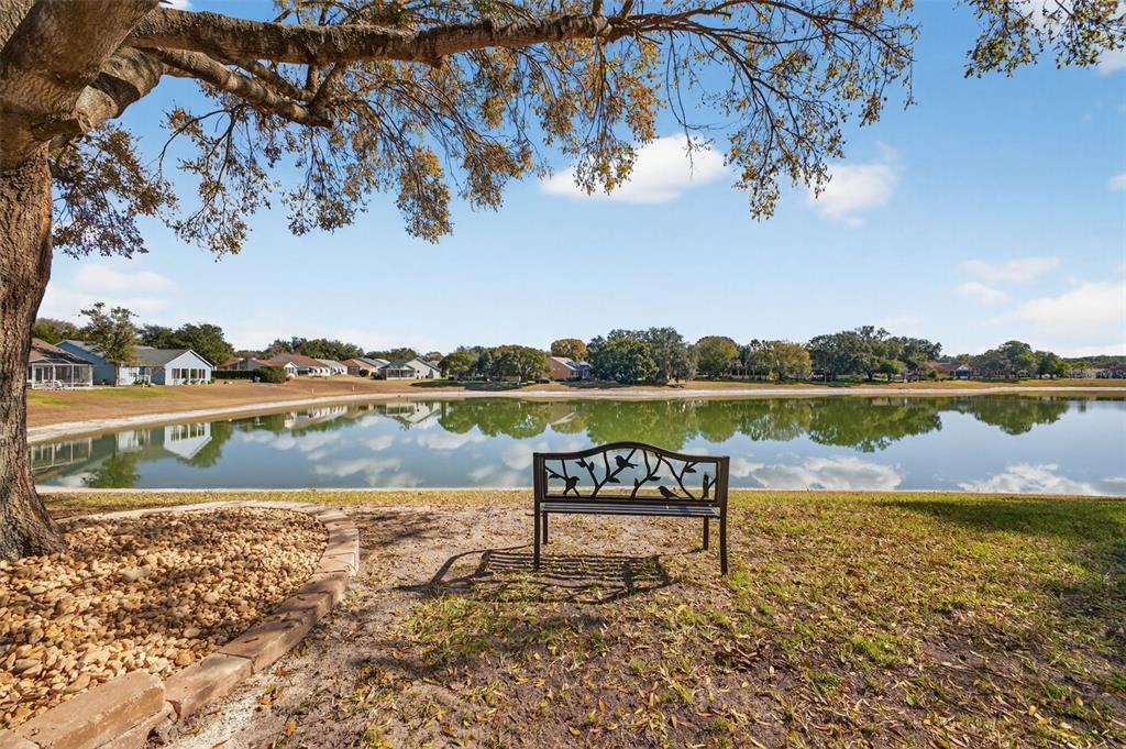 A metal bench sits on a grassy lawn overlooking a calm lake with distant houses — 411 DEL MAR DRIVE, LADY LAKE, FL