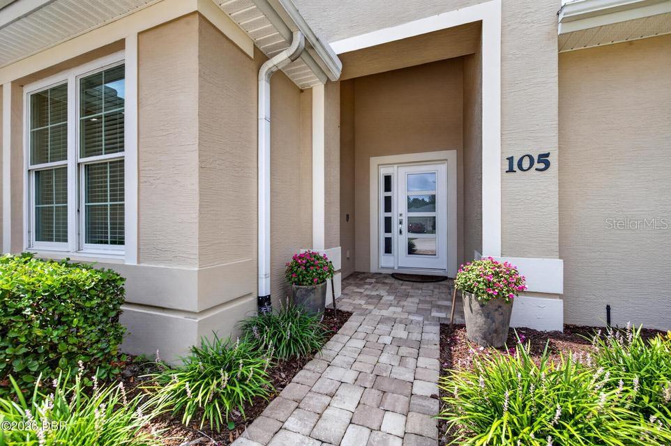 A paved walkway leads to the front entrance of a beige house with a white door — 105 OPAL HILL CIRCLE, DAYTONA BEACH, FL