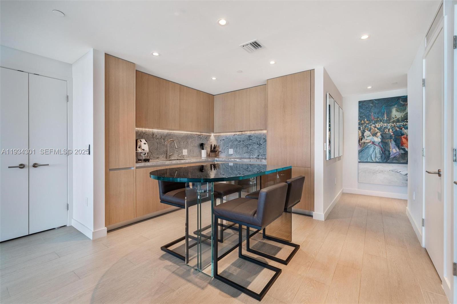 A modern kitchen features light wood cabinetry, a glass-topped island, and dark leather bar stools — 500 Alton Rd # 1602, Miami Beach FL 33139, MIAMI BEACH, FL