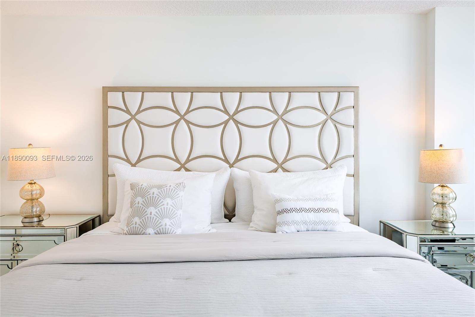 A bedroom features a white bed with a geometric headboard and mirrored nightstands with lamps — 6917 Collins Ave # 720, Miami Beach FL 33141, MIAMI BEACH, FL
