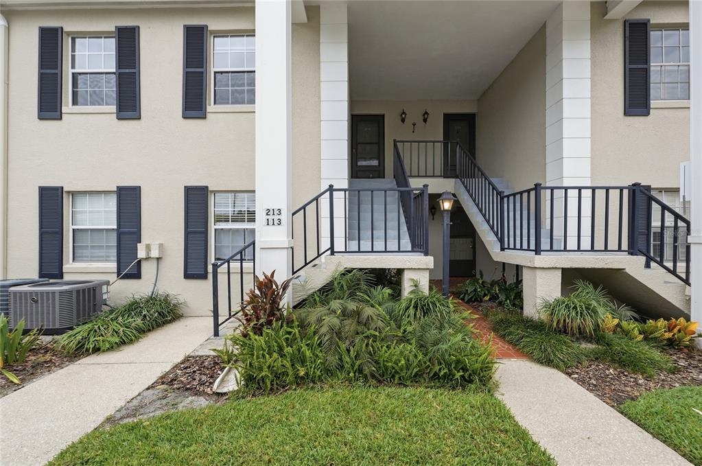 A two-story apartment building exterior features a shared staircase, dark railings, and landscaped front gardens — 689 LAKE HOWARD DRIVE NW UNIT 213, WINTER HAVEN, FL