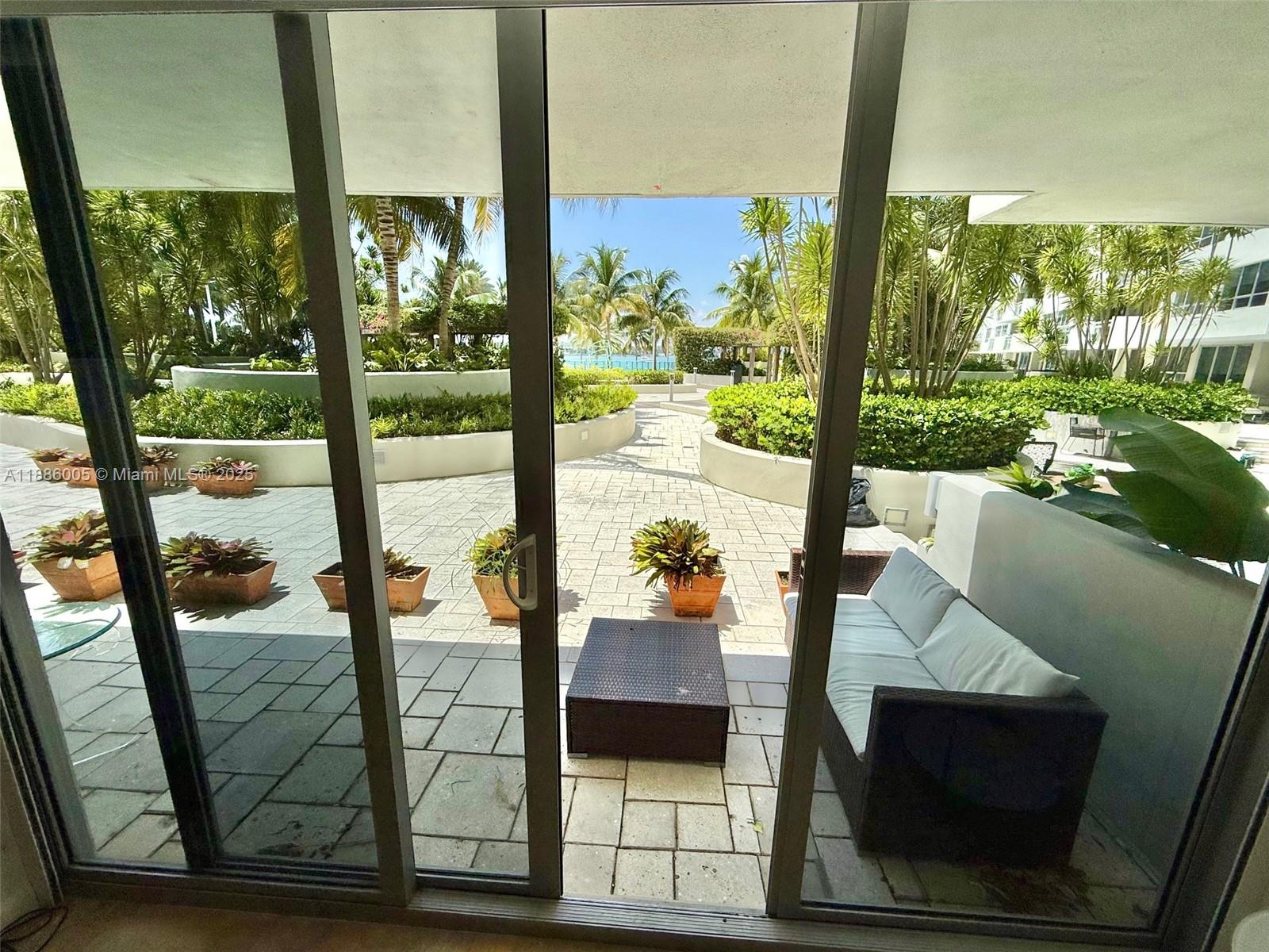A patio with a sofa and potted plants overlooks a landscaped courtyard with palm trees — 1500 Bay Rd # 122S, Miami Beach FL 33139, MIAMI BEACH, FL