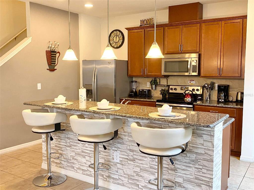 A kitchen features a granite-topped breakfast bar, white stools, wooden cabinets, and stainless steel appliances — 3175 TOCOA CIRCLE, KISSIMMEE, FL