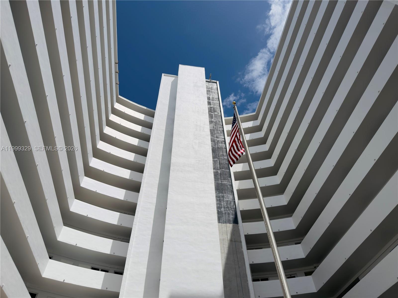 A low-angle view of a white multi-story building with balconies and an American flag — 710 N Ocean Blvd # 603, Pompano Beach FL 33062, POMPANO BEACH, FL