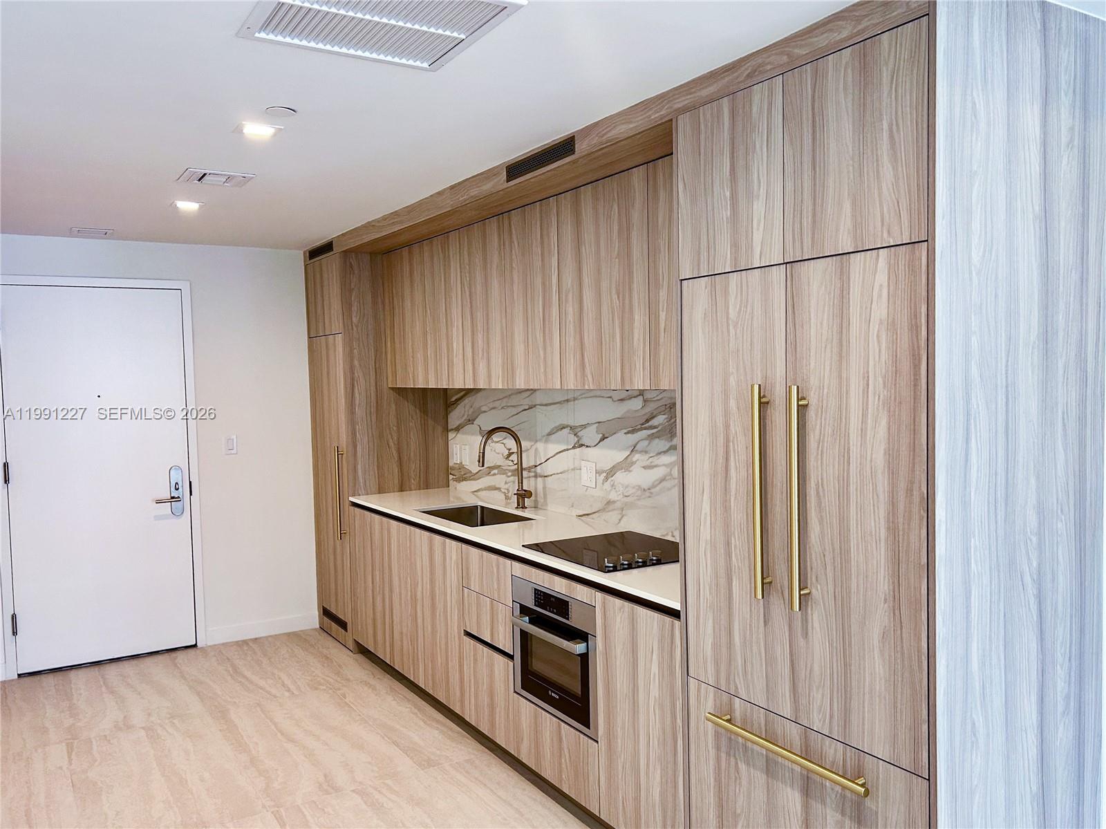 A modern kitchen features wood-grain cabinetry, a marble backsplash, and gold hardware — 77 SE 5TH ST. # 3614, Miami FL 33131, MIAMI, FL